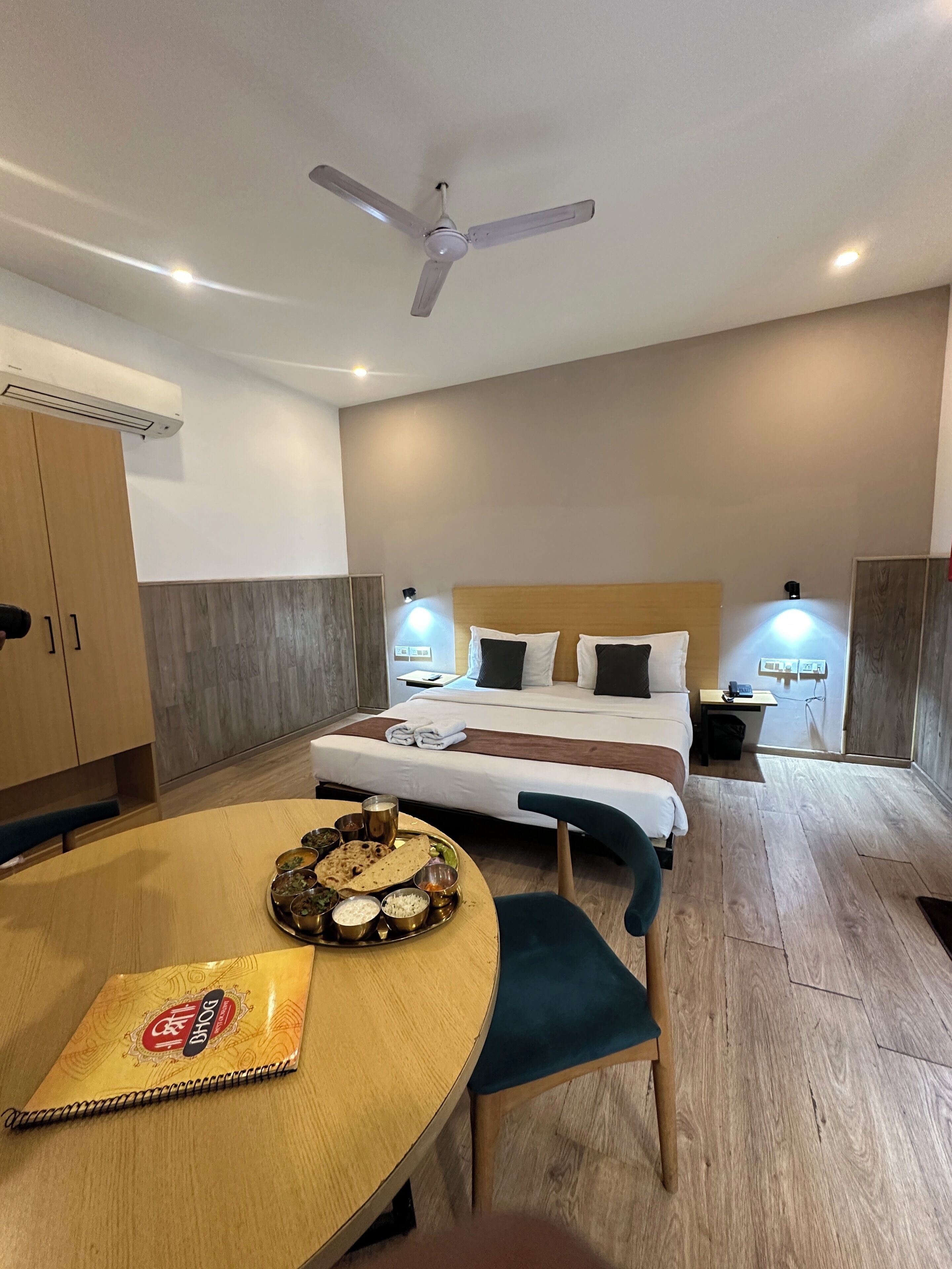 A hotel room with a bed, bedside tables, a desk, a chair, a dining table with food, and a ceiling fan.