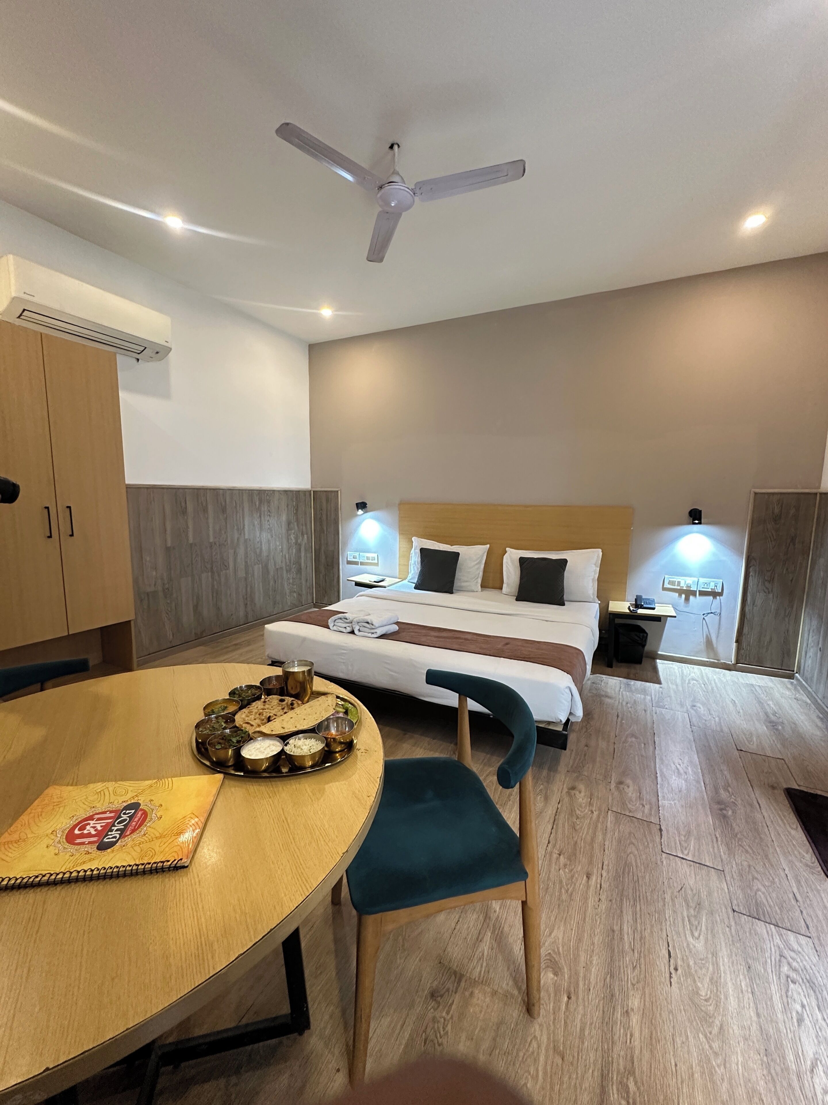 Une chambre d’hôtel comprenant un lit, des tables de chevet, un ventilateur de plafond, un appareil de climatisation, une table à manger avec une assiette de nourriture et une chaise.