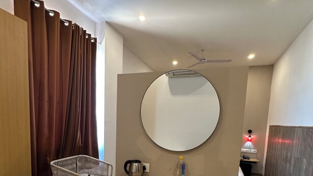 Une chambre d’hôtel avec un plancher en bois, un miroir rond, une petite table avec une bouilloire, une chaise et un lit à l’arrière-plan.