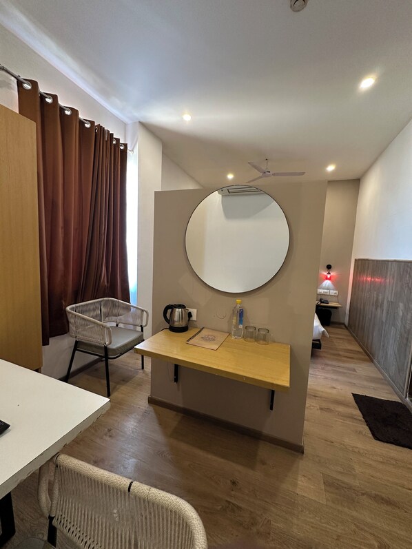 Une chambre d’hôtel avec un plancher en bois, un miroir rond, une petite table avec une bouilloire, une chaise et un lit à l’arrière-plan.