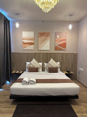 A modern hotel room with a large bed, wooden headboard, three wall art pieces, two bedside tables with phones, and a chandelier.