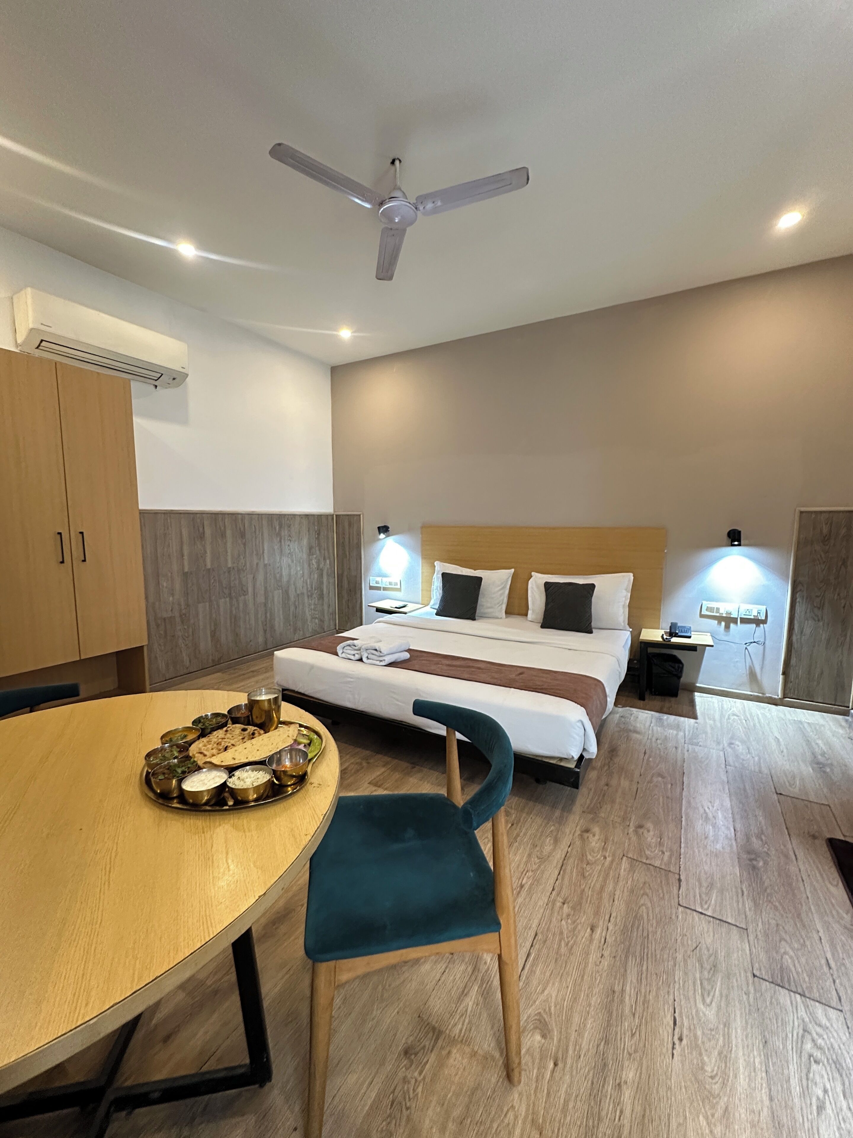 A hotel room with a bed, bedside lamps, a ceiling fan, an air conditioning unit, a dining table with a tray of food, and a chair.