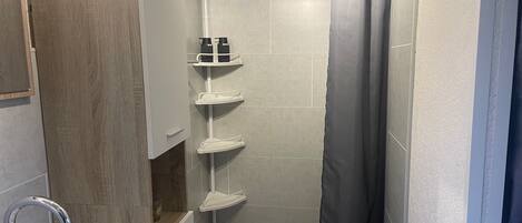 Un baño con ducha, lavabo e inodoro. Hay estantes con artículos de aseo.