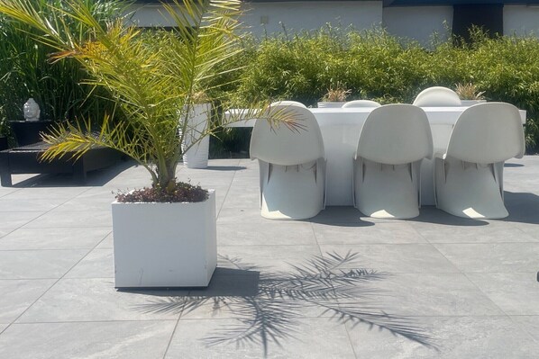 Eine Terrasse mit einem weißen Tisch und Stühlen, umgeben von Topfpflanzen und einem gefliesten Boden.