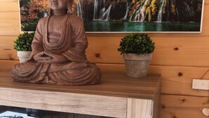 Une statue en bois représentant une figure assise, deux plantes en pot et un tableau encadré sur un mur en bois.