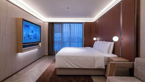 Ein modernes Hotelzimmer mit einem großen Bett, einem flachbildfernseher an der Wand, einem Nachttisch mit Lampe und Blick auf die Stadt durch große Fenster.
