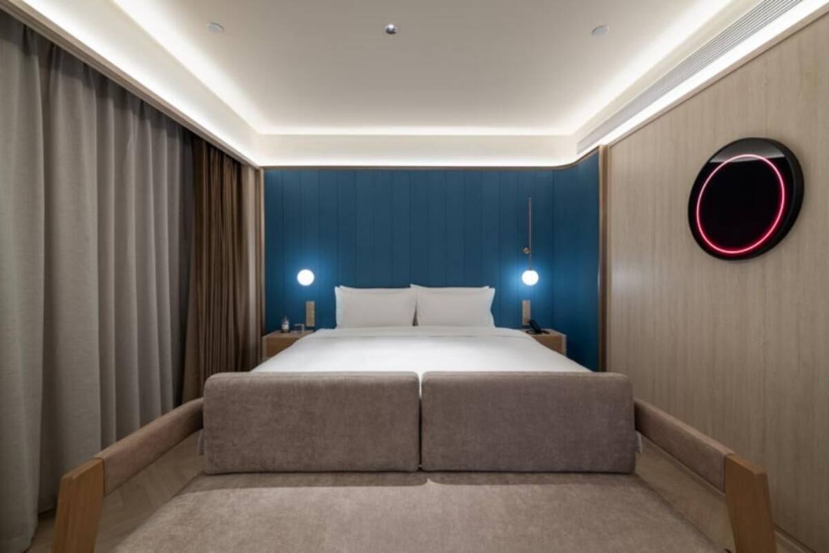 Moderni hotellihuone, jossa on suuri sänky, yöpöytävalaisimet, pyöreä kattovalaisin ja puinen tehosteseinä.