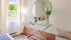 Een slaapkamer met een glazen tafel, een spiegel, een vaas met bloemen, een bed en uitzicht op groen.
