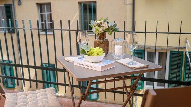 Een tafel met een boek getiteld 'Henri Matisse', een kom groene druiven, een vaas met bloemen en twee glazen op een balkon.