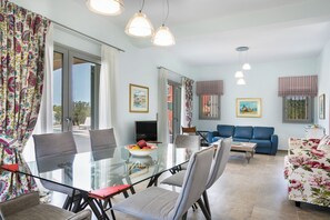 A modern living room with a glass dining table, blue sofa, and a TV. - Afrodite Luxury Villa, Estrellas Luxury Villas (Kefalonia)