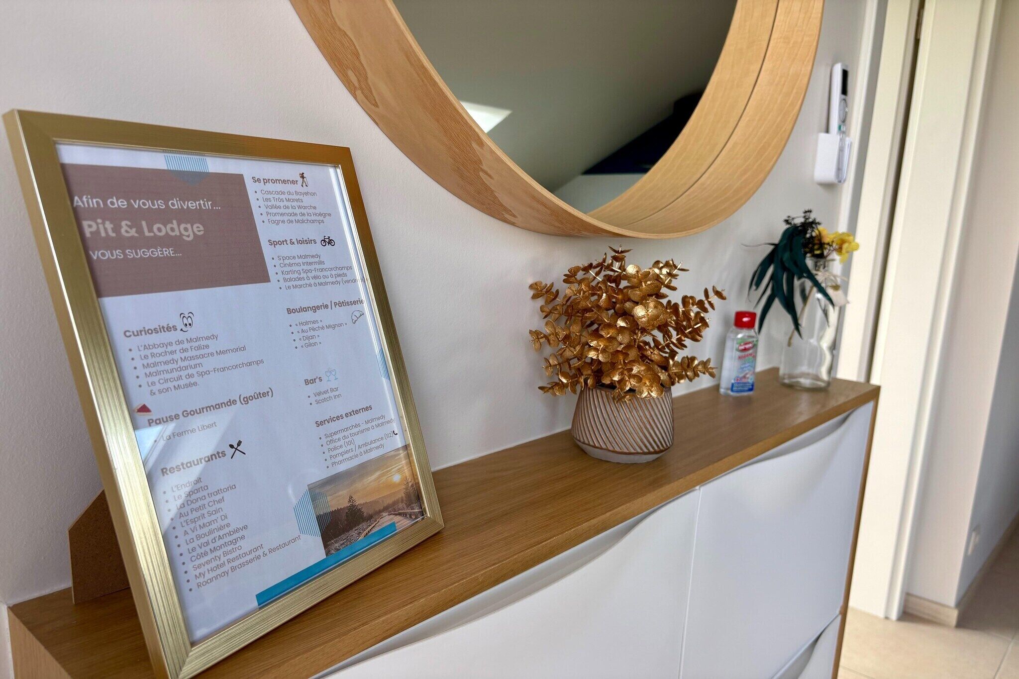 Un panneau d’information encadré est posé sur un comptoir en bois, au-dessus duquel se trouve un miroir. On y voit également un vase décoratif orné de feuilles dorées et une petite plante en pot.