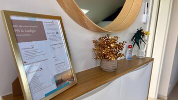 Un panneau d’information encadré est posé sur un comptoir en bois, au-dessus duquel se trouve un miroir. On y voit également un vase décoratif orné de feuilles dorées et une petite plante en pot.