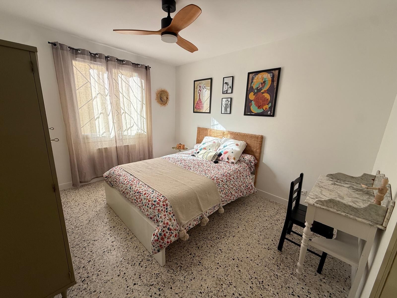 Une chambre à coucher comprenant un lit, un bureau, une chaise, un ventilateur de plafond et des œuvres d’art accrochées au mur.