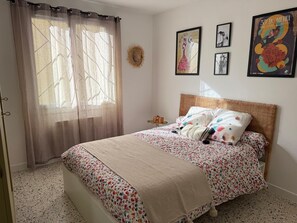 Ein Schlafzimmer mit Bett, einem Fenster mit leichtem Vorhang, einem gewobenen Kopfteil und Wandbildern.
