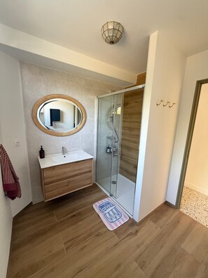 A bathroom with a wooden vanity, a round mirror, a glass shower enclosure, and a door with a towel rack. - P2 camarguais calme avec terrasse en centre ville (Aigues-Mortes)
