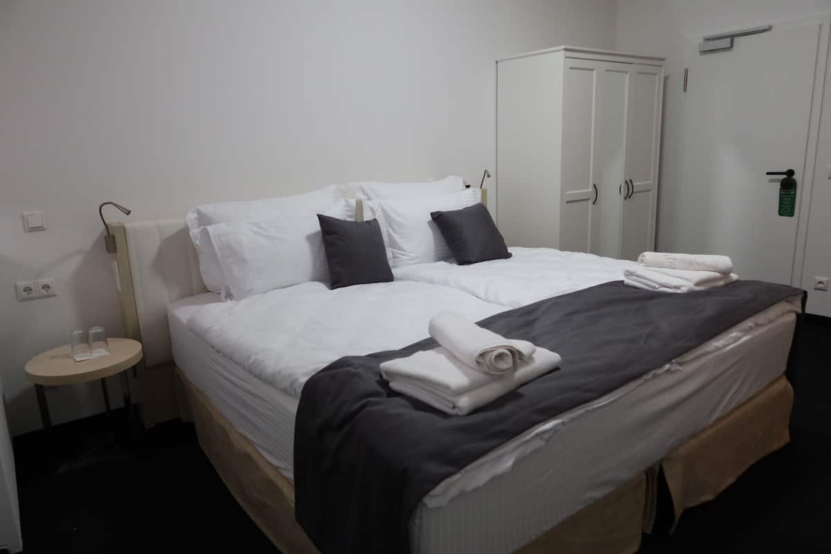A neatly made bed with white and dark pillows, a folded towel, and a dark blanket. There is a small bedside table with a lamp and a white wall socket. A white wardrobe is visible in the background.