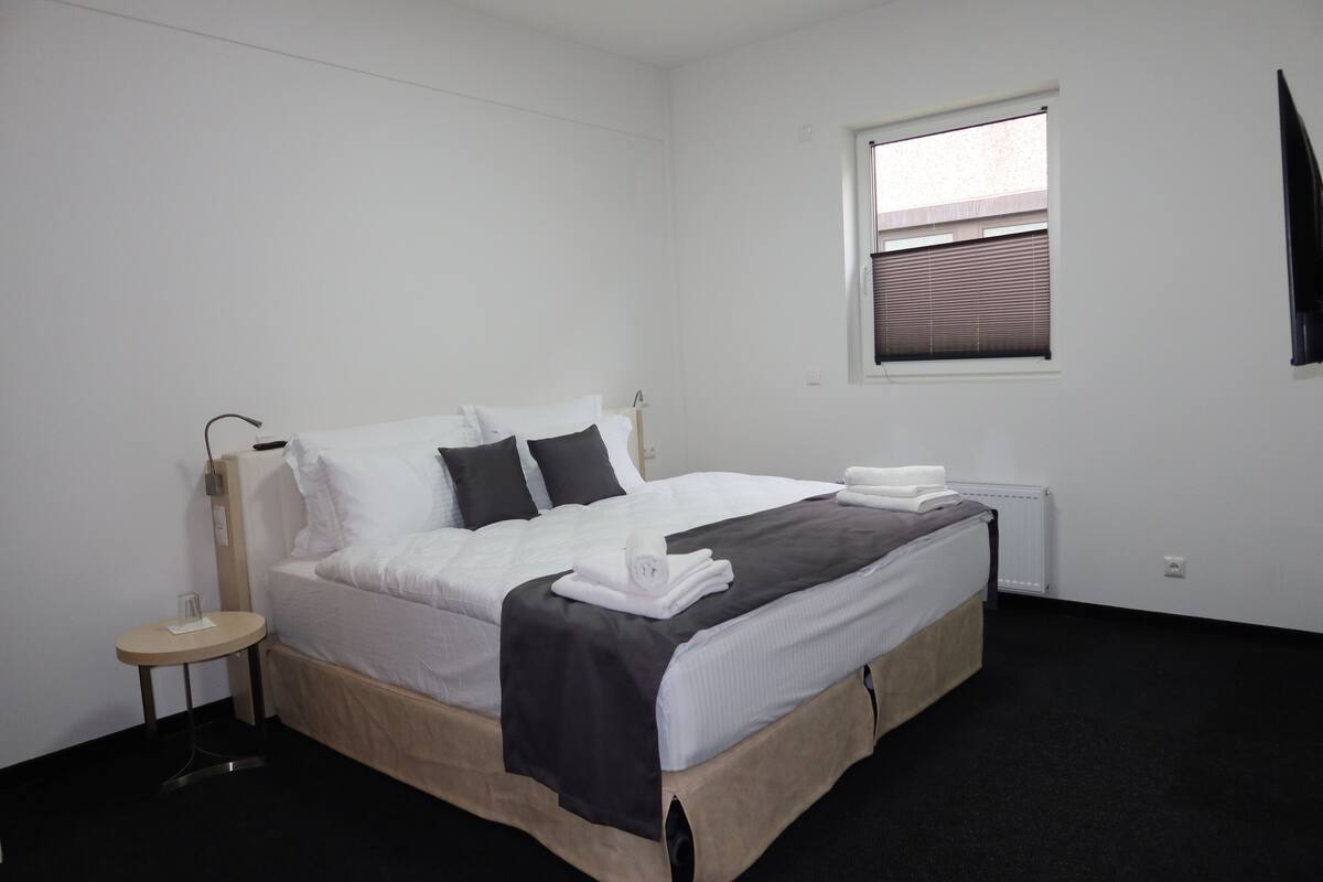 A single bed with a black and white duvet, a bedside table with a lamp, and a window with blinds.