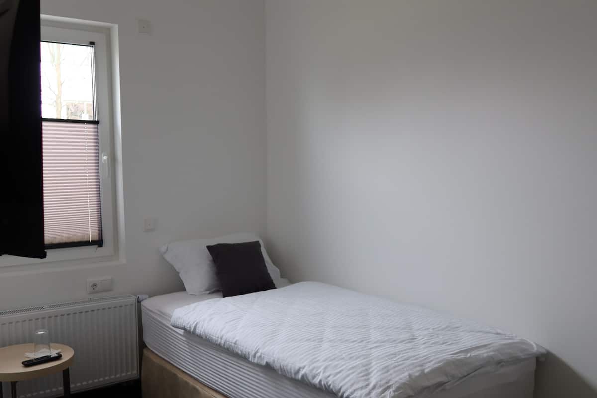 A small, single-bed room with a window, a radiator, a small table, and a stool.