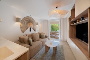A modern living room with a sofa, a round coffee table, a TV mounted on the wall, and a large window with a view of greenery outside. - 6P apartment in a Citadelle /POOL - Saint-Tropez (Saint-Tropez)