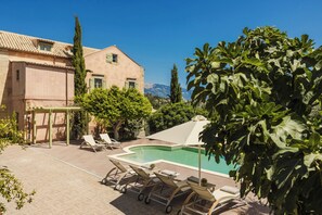 Ein Poolbereich mit Liegestühlen und einer Pergola, umgeben von Bäumen und einem Gebäude mit Ziegeldach.