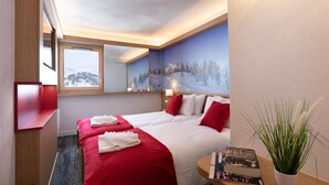 Hotelkamer met twee bedden, een groot raam, een schilderij van een besneeuwd landschap en een tafeltje met een plant.
