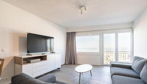 A modern living room with a flat-screen TV, a sofa, a coffee table, and a balcony with a view.