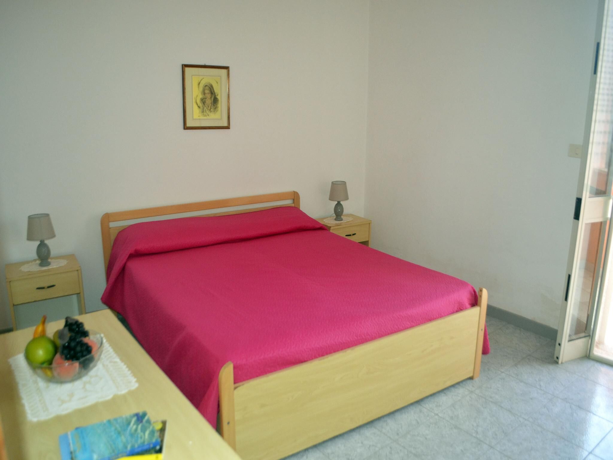 A bedroom with a wooden bed, two bedside tables with lamps, a framed picture on the wall, and a small table with fruit.