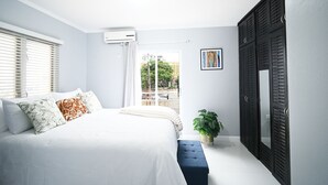 Ein Schlafzimmer mit Bett, einem Fenster mit Jalousien, einer Zimmerpflanze und einer Tür mit Fensterläden.