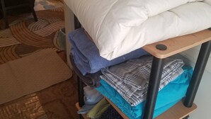 A neatly made bed with a white comforter and pillows, a wooden shelf with folded clothes, and a patterned rug on the floor. - Adorable Tiny House Near Mercedes Benz Stadium (Atlanta)