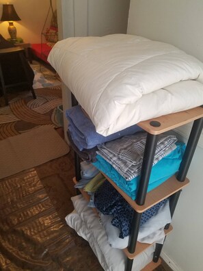 A neatly made bed with a white comforter and pillows, a wooden shelf with folded clothes, and a patterned rug on the floor. - Adorable Tiny House Near Mercedes Benz Stadium (Atlanta)