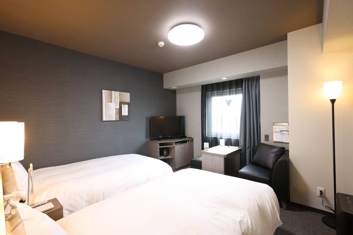 A hotel room with two beds, a desk, a chair, a television, and a lamp.
