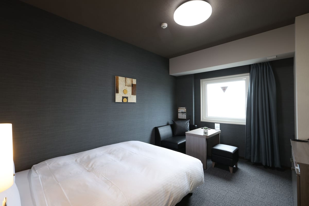 A hotel room with a bed, a desk, a chair, a small table, a window with curtains, and a wall-mounted artwork.