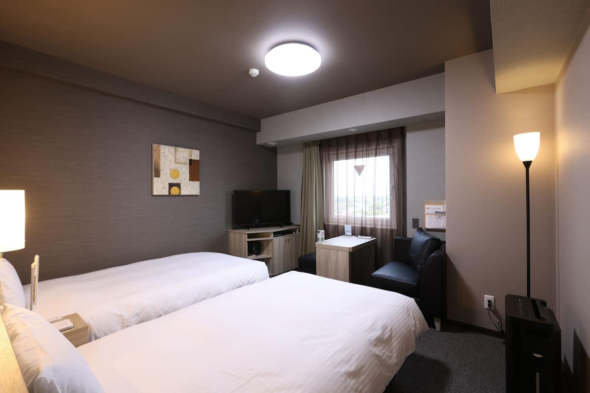 A hotel room with two beds, a desk, a chair, a television, and a lamp.