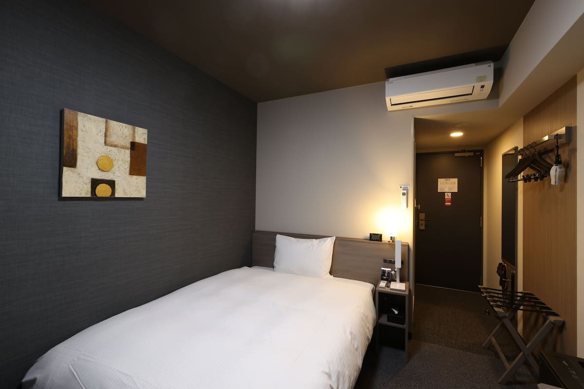 A hotel room with a bed, a nightstand, an air conditioning unit, a coat rack, and a door leading to another room.