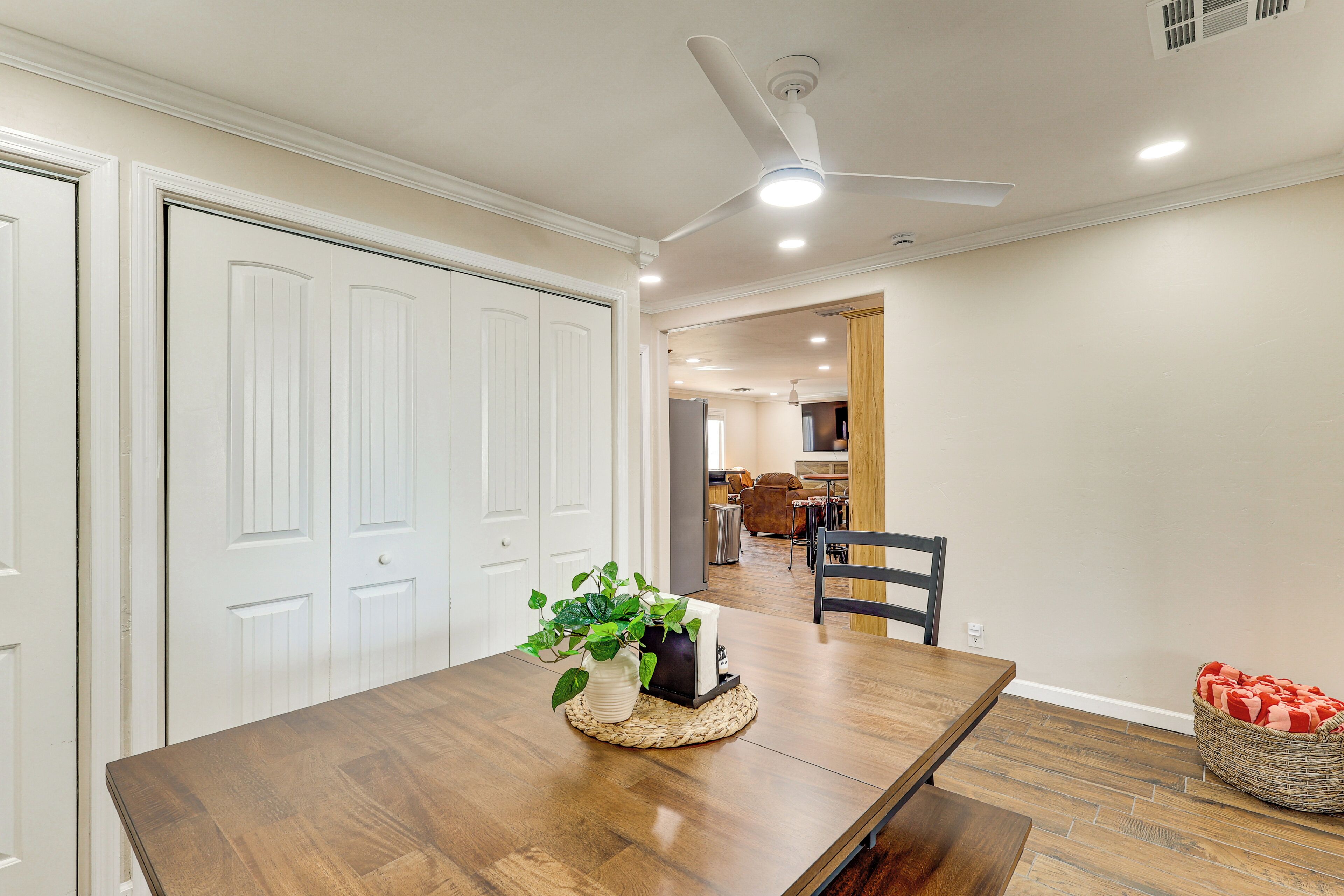 Un coin repas avec une table en bois, des chaises, un ventilateur de plafond et, au fond, une cuisine.