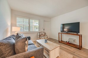 A living room with a sofa, armchair, coffee table, TV stand, and a window with blinds. - Poolside Fun, Ocean Strolls! Naples Beach Getaway (Naples)