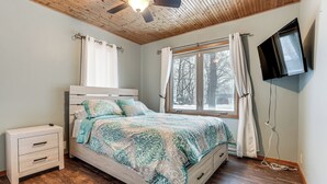 A bedroom with a bed, a nightstand, a ceiling fan, a window with curtains, and a TV mounted on the wall. - Lakefront Home w/ Views, 7 Mi to Dtwn Paw Paw (Paw Paw)