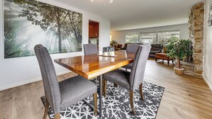 A dining area with a wooden table, gray upholstered chairs, a large forest-themed painting, and a stone accent wall. - Lake View! South End Seattle Home Near Lumen Field (Seattle)