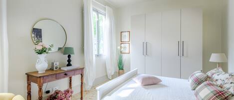 Une chambre à coucher comprenant un lit, des tables de chevet, une commode, un miroir et une fenêtre avec des rideaux.