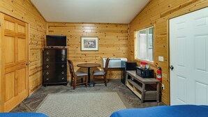 A small, cozy room with wooden walls, a wooden dresser, a round table with chairs, a TV, a framed picture, and a window with blinds.