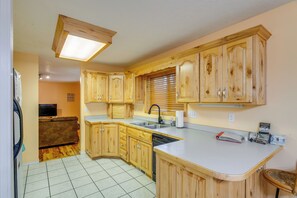 A kitchen with wooden cabinets, a built-in dishwasher, and a window with blinds. - Extended-Stay 'Geyser Home' Near Yellowstone NP (West Yellowstone)