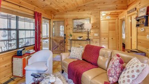 Interior de una cabaña de madera con un sofá de cuero, un sillón blanco, una mesita con una lámpara y un cuadro en la pared.