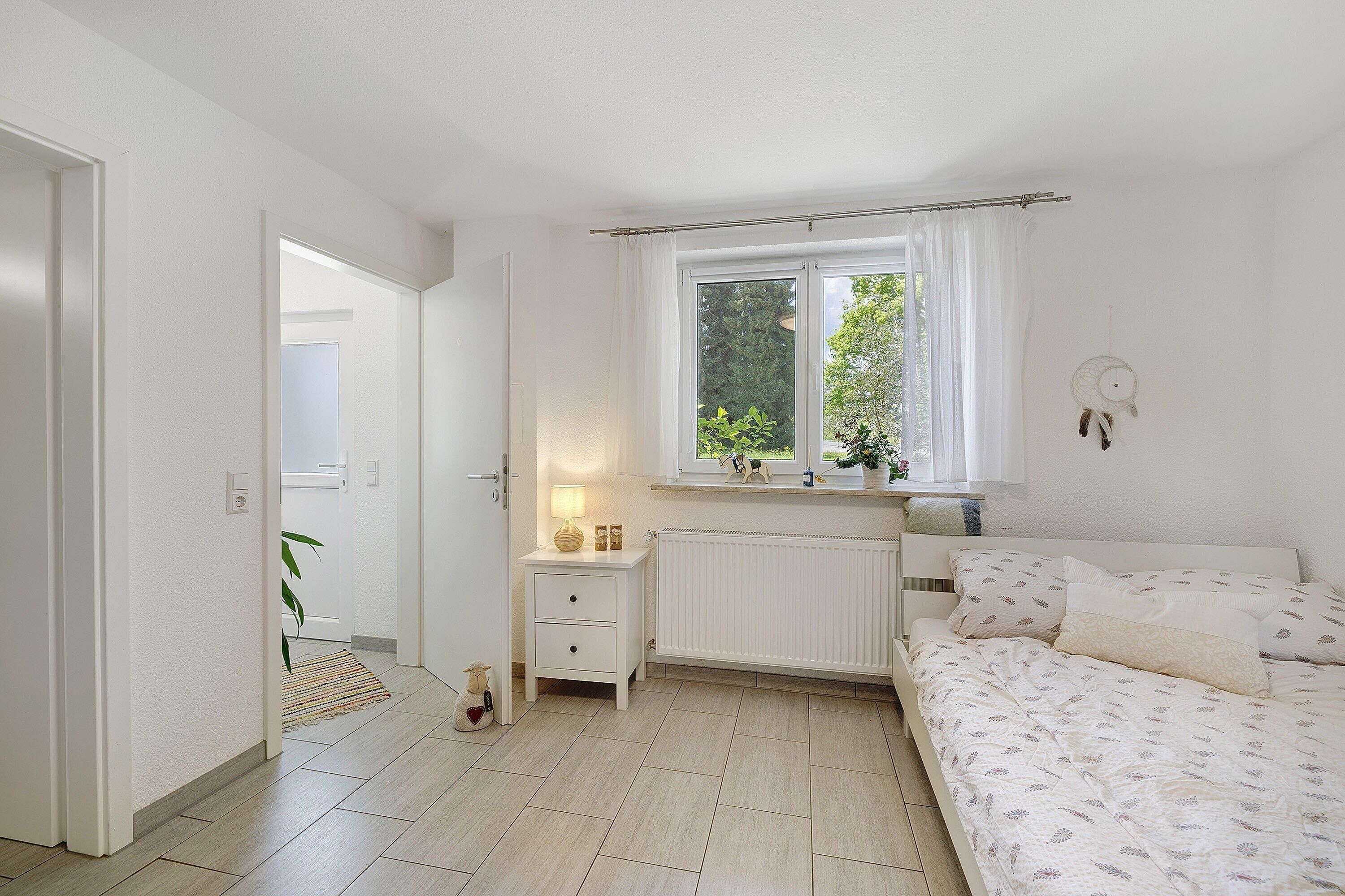 Un dormitorio con cama, mesita de noche, ventana con cortinas, radiador y puerta que da a otra habitación.