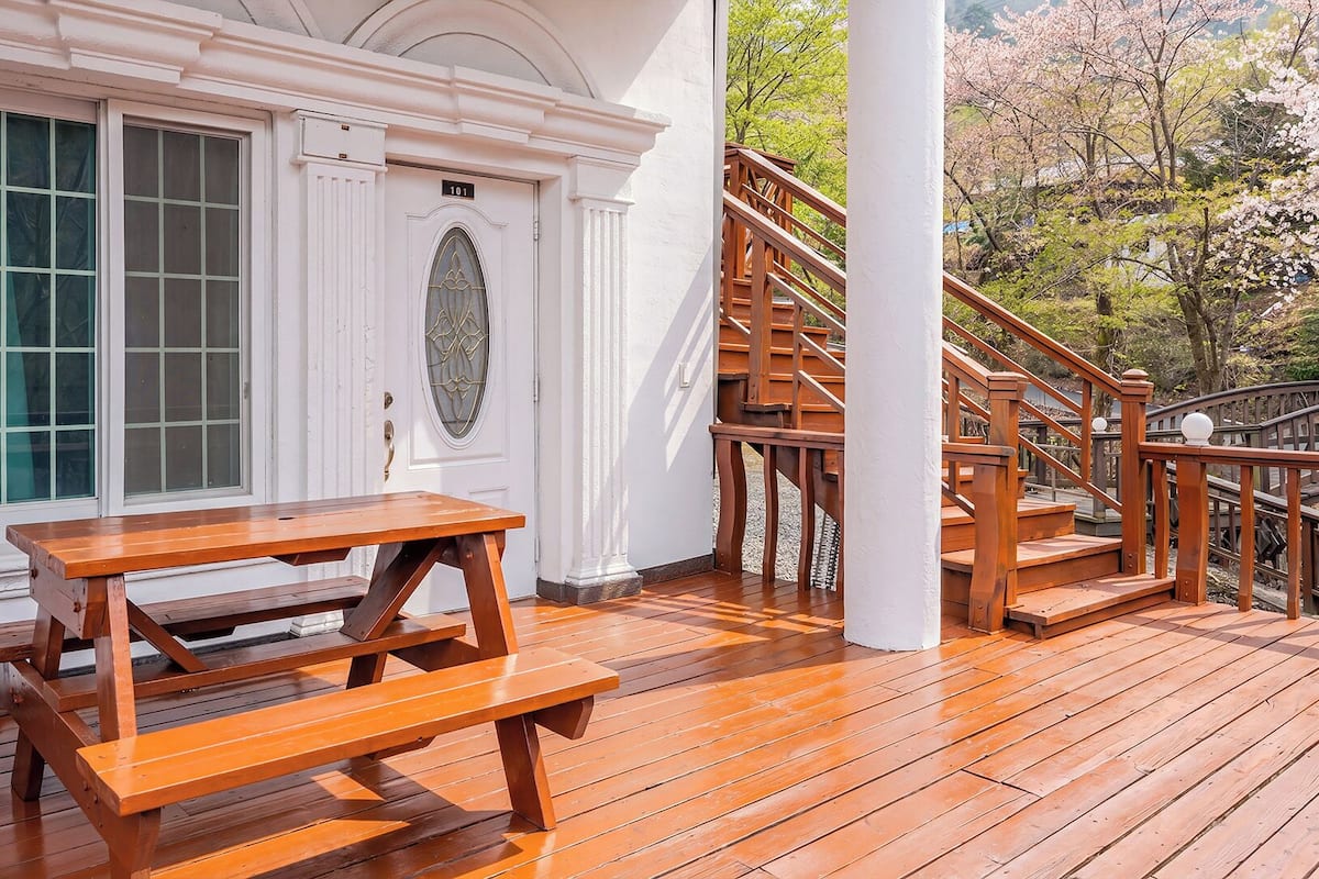 Una terraza de madera con una mesa de picnic, un edificio blanco con un cartel del número 101 y una escalera que sube a un nivel superior.
