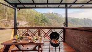 Un área de comedor al aire libre con una mesa de madera dispuesta para una comida, una parrilla con alimentos y vistas a un paisaje boscoso.