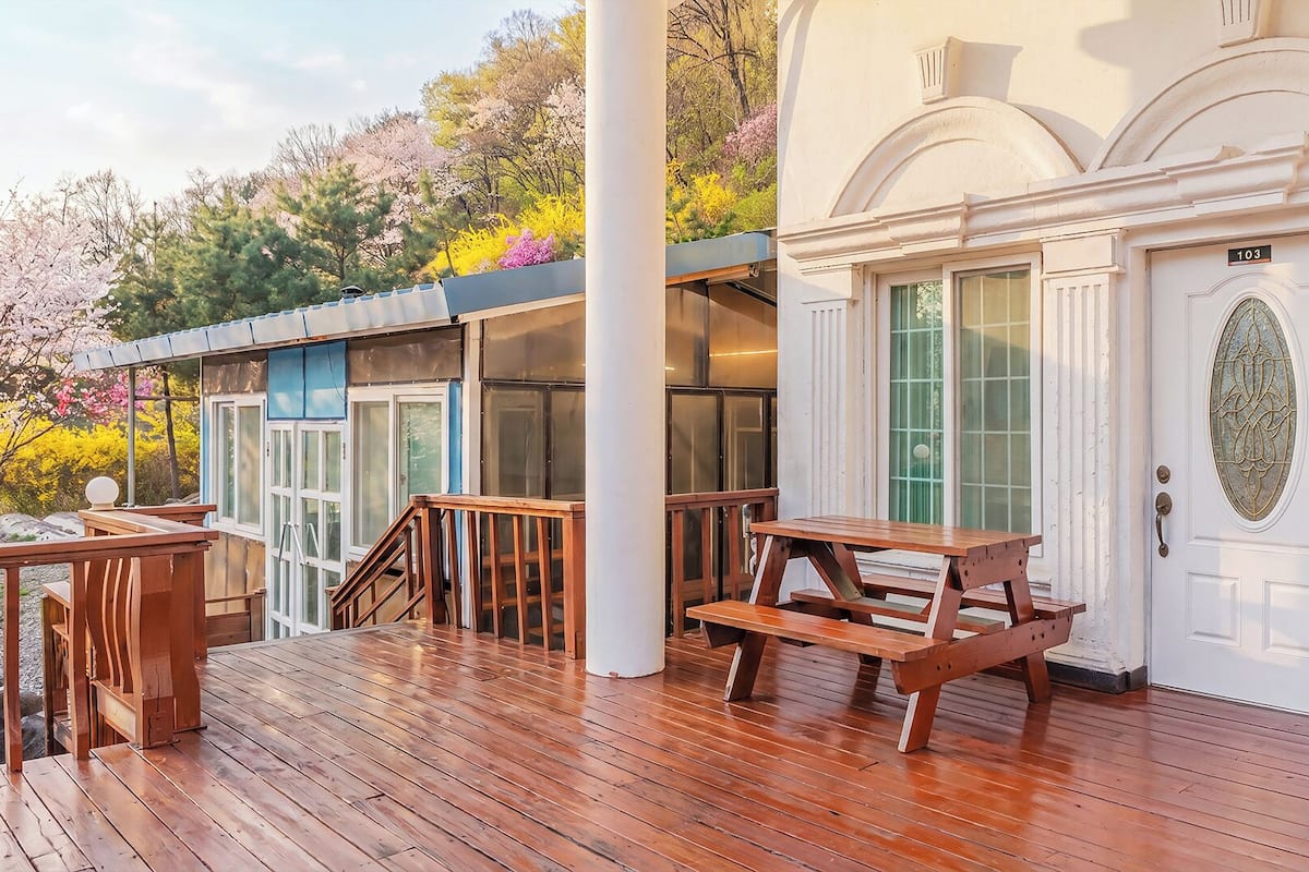 Una terraza de madera con una mesa de picnic, un edificio blanco con una puerta y una ventana con un panel de vidrio decorativo.