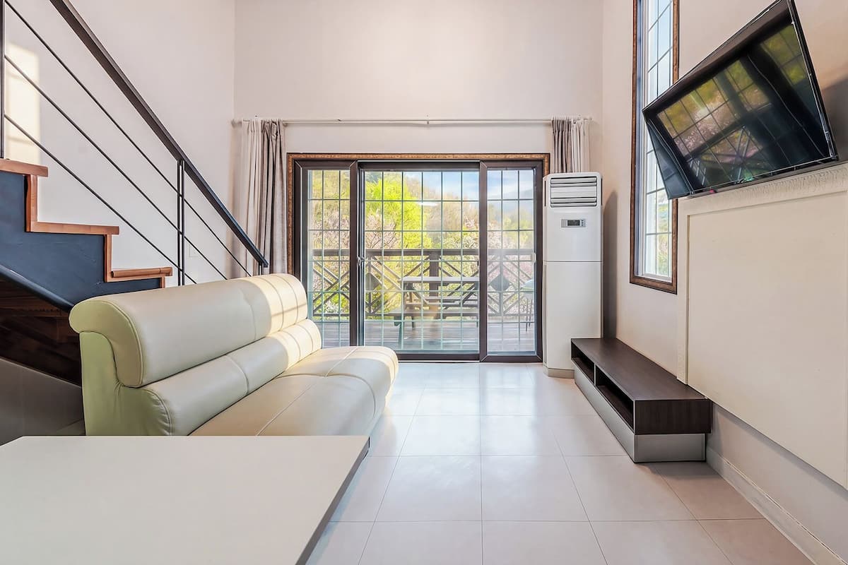 Una sala de estar moderna con un sofá, un televisor montado en la pared, una escalera y un amplio ventanal corredera que da a un espacio exterior.