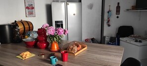 Eine Küche mit Kühlschrank, einem Holzfass, einem Wasserspender, einer Vase mit Blumen, einem roten Wasserkocher, einem Holzschnittbrett, einer Platte mit Brot sowie einer blauen und einer roten Tasse.