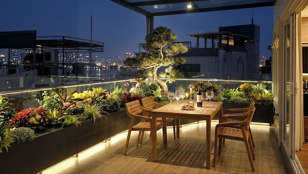 Terraza en la azotea con cerramiento de vidrio, mesa de madera, sillas, plantas en macetas y vistas a la ciudad al atardecer.