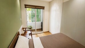 Ein Schlafzimmer mit Bett, Stuhl, kleinem Tisch, einem Fenster mit Jalousien und Blick ins Freie.